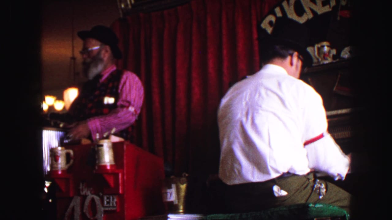 1968 Vancouver 8mm frame of a dim saloon interior with two adult men in hats, red curtains, a counter, and visible barware.