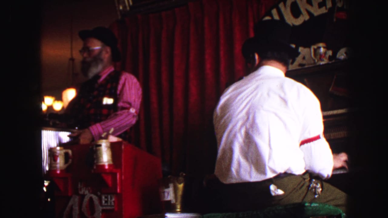 Dim stage interior with a man at a piano and another man behind a red counter.