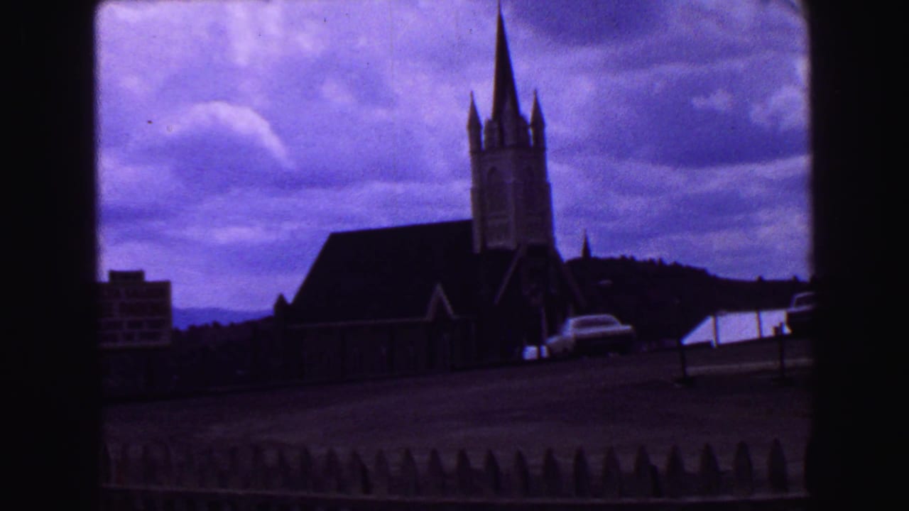Church with tall steeple, cloudy sky, parked car, and fence in foreground.