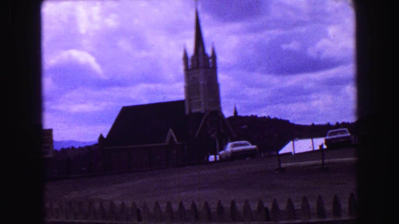 Steepled church and parked car viewed across a sloping road under clouds.
