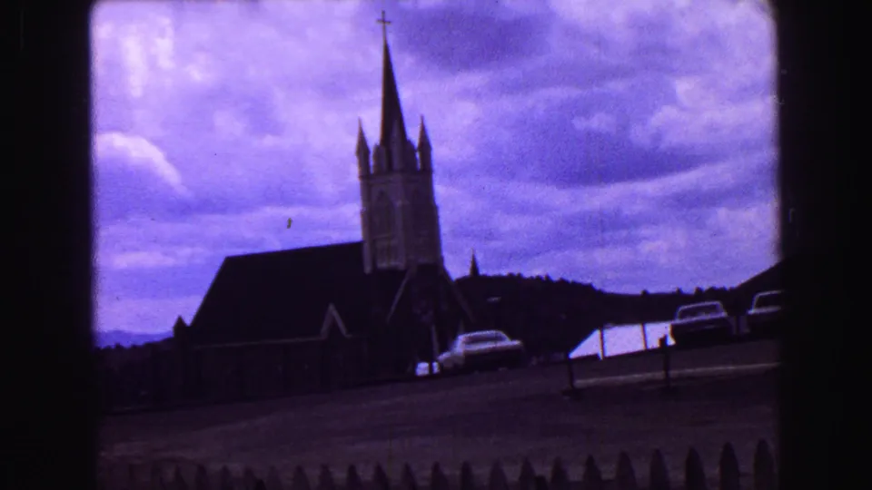 1968 Vancouver frame with a tall church steeple, dark roof, roadside parking, picket fence, and distant hills.