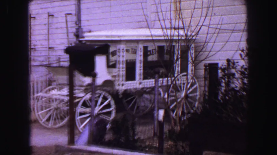 1968 Vancouver home-movie frame showing a white carriage near a building, posts, fence, shrubs, and bare tree branches.