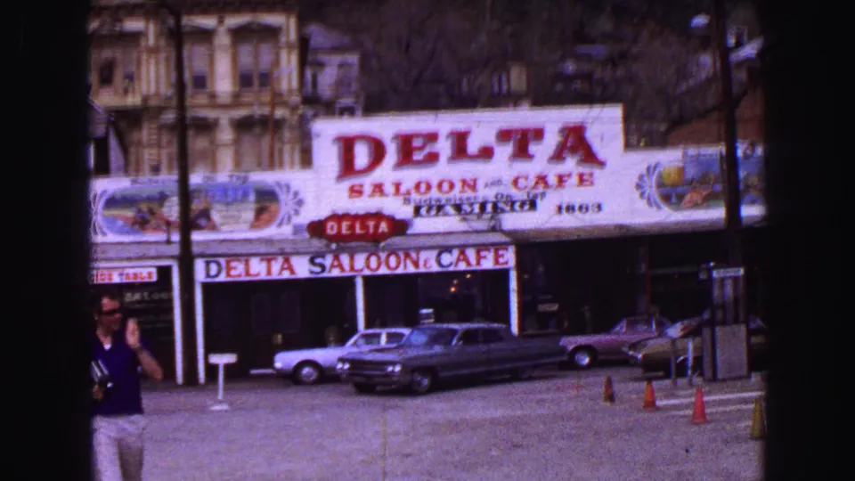 1968 Vancouver frame showing the Delta Saloon and Cafe facade, red signage, parked 1960s cars, cones, and a pedestrian near frame edge.