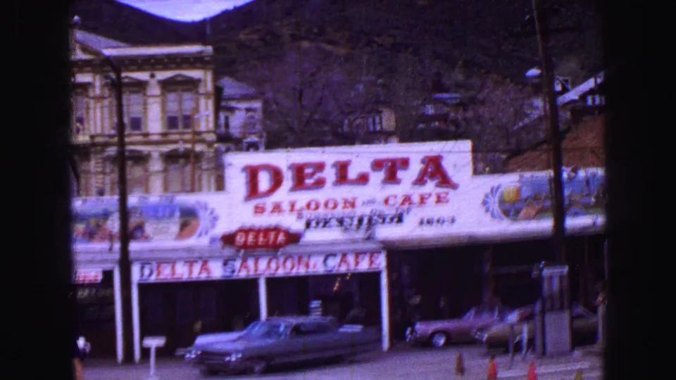 1968 8mm frame of the Delta Saloon & Cafe storefront, parked cars, utility poles, and dark hillside in the background.