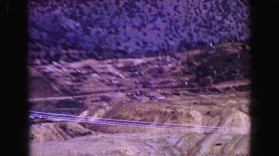 Vintage 8mm frame with dry roadside terrain, utility lines, scrub vegetation, and dark gate edges during a moving landscape shot.