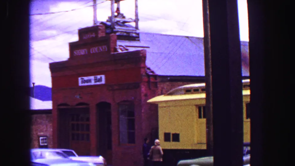 1968 home-movie frame of a brick building beside a yellow rail car, with a parked car and small pedestrians visible through window framing.