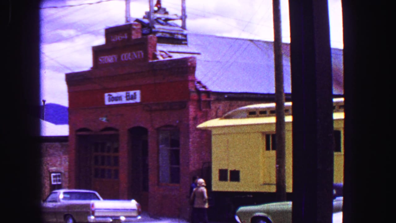 Town-hall facade and yellow rail car seen through dark vertical obstructions, with a parked blue car.