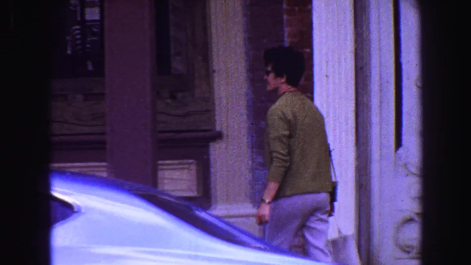 1968 Vancouver-area 8mm frame showing a person in a green sweater walking past storefront architecture and the hood of a parked car.