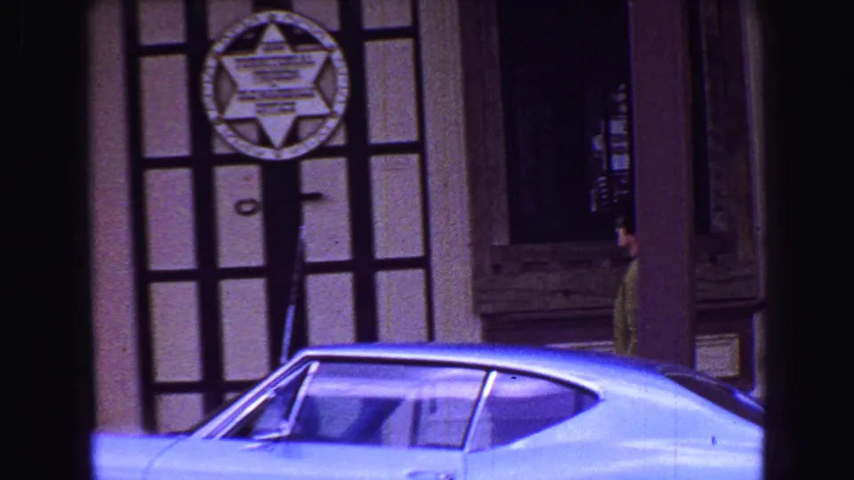 1968 Vancouver-area street frame with a sheriff-star door emblem, parked blue car, and a person walking along the building frontage.