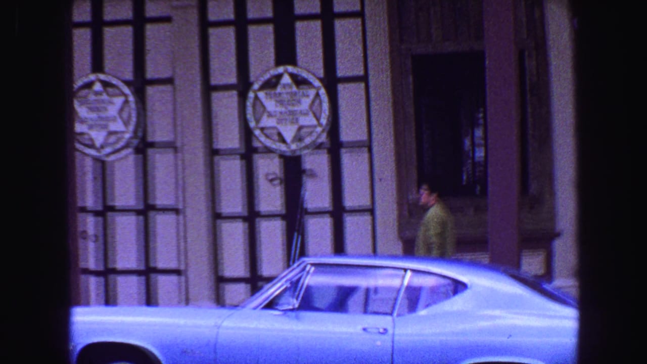 1968 Vancouver home-movie frame showing a light blue coupe outside a barred Territorial Prison facade with sheriff badge signs.