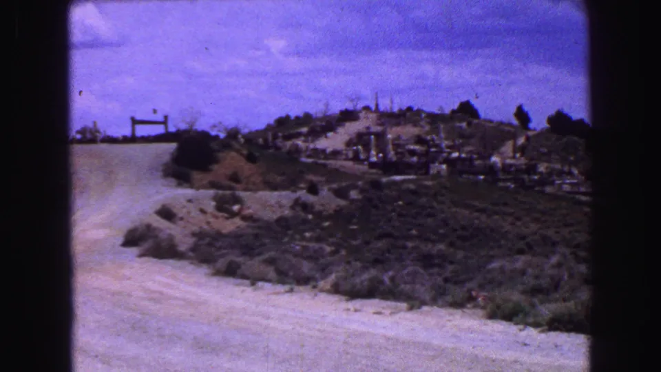 1968 home-movie view of a dusty unpaved road leading toward a scrubby hillside cemetery under a hazy sky.