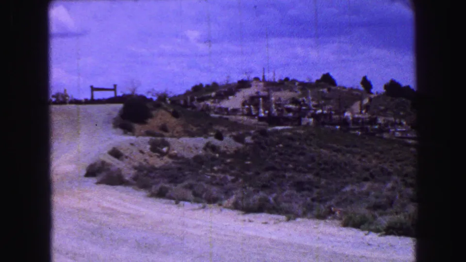 Late-1960s 8mm frame of a pale dirt road, scrub-covered hillside, and scattered cemetery markers in Vancouver footage.