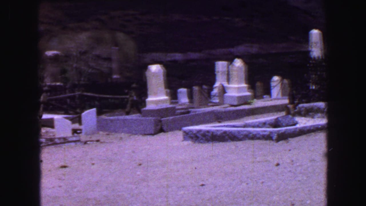 White headstones and low grave borders in a cemetery beside a rocky hillside.