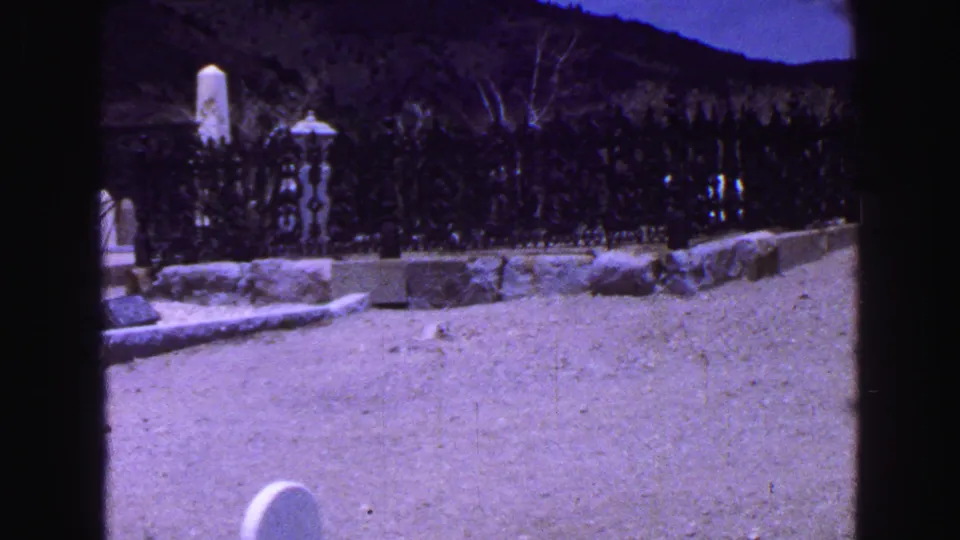 At the end of the 1968 Vancouver cemetery pan, dark fencing, shrubs, a white marker, and open pale ground fill the frame.