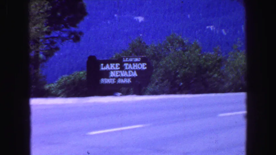 Roadside 8mm frame showing a readable sign for leaving Lake Tahoe Nevada State Park, with trees, mountains, blue sky, and pavement in view.