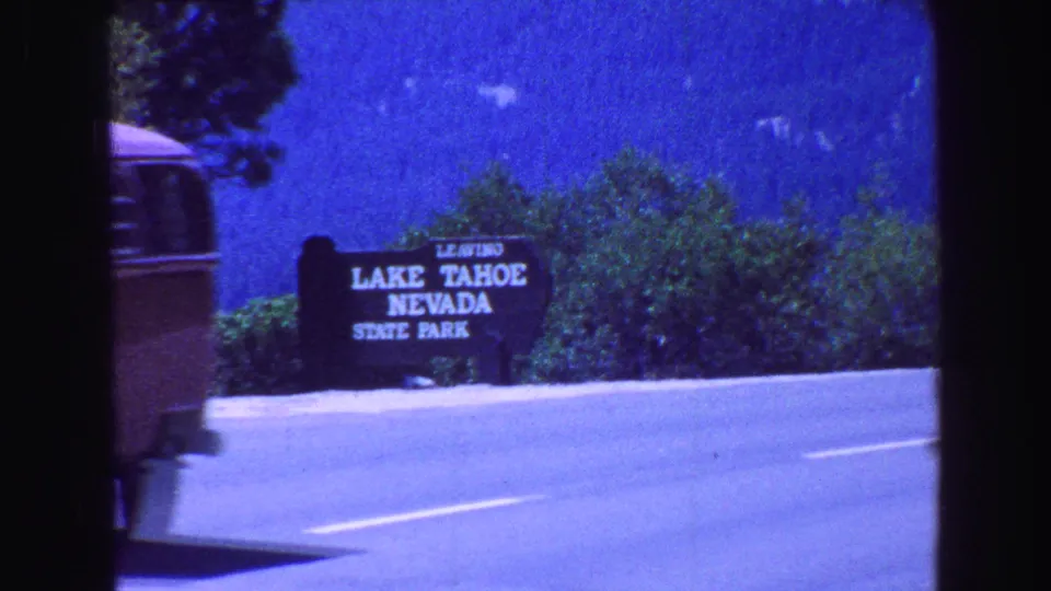 The Lake Tahoe Nevada State Park sign stands beside the highway with trees and bright blue sky behind it.