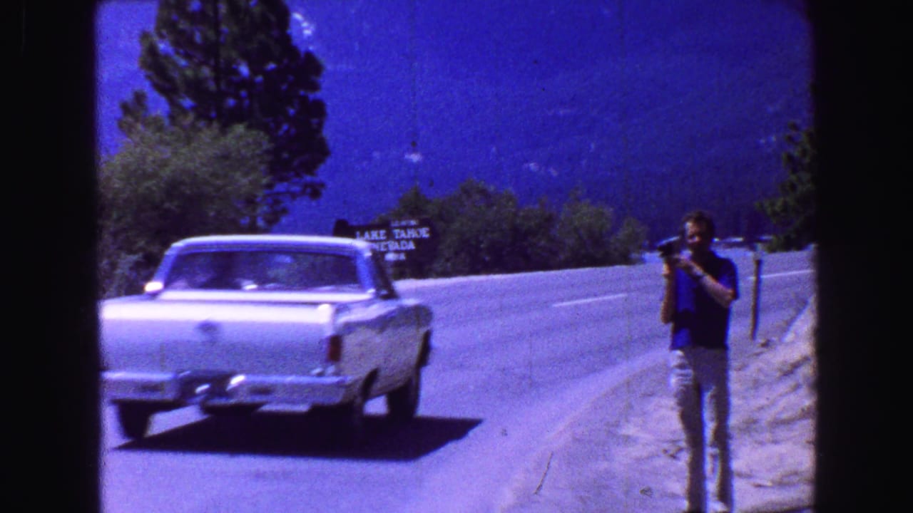 1960s 8mm frame of a white car passing an adult man with a camera beside a mountain road, with a roadside sign and wooded slopes behind.