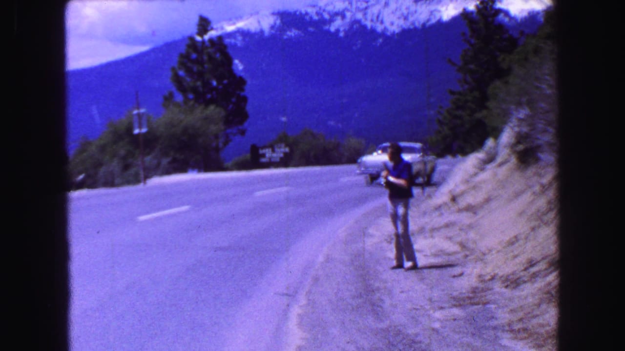 Man standing on the shoulder of a winding mountain road with cars in the distance.
