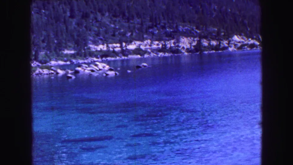 Handheld 8mm view of vivid blue lake water with submerged dark patches, pale rocks along the shore, and dense evergreens rising behind.