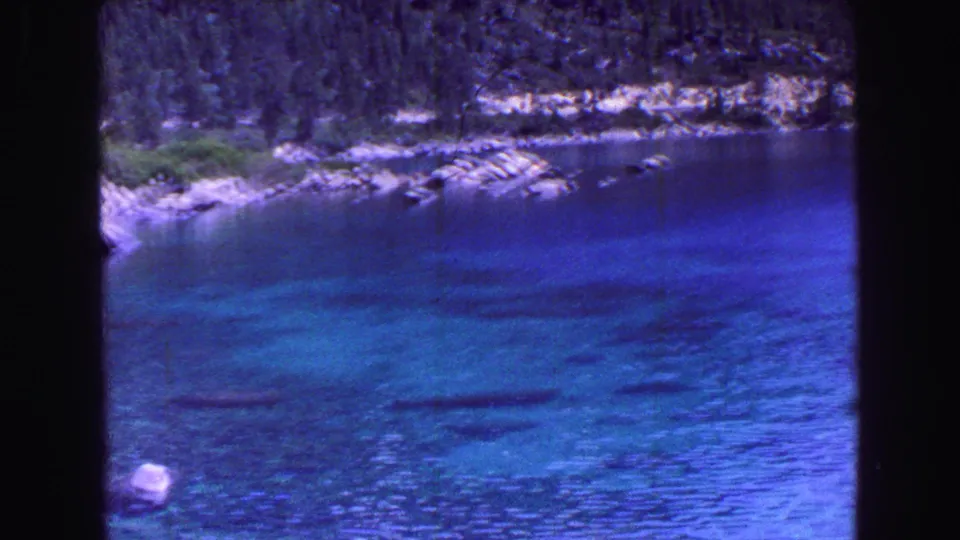 Blue 8mm frame of clear lake water near a rocky shore, with evergreen-covered hills and pale stones visible along the opposite bank.