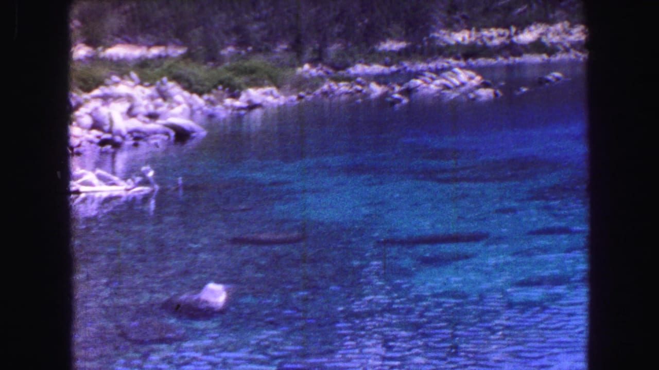 Clear blue water with submerged stones, shoreline rocks, foliage, and dark film borders.