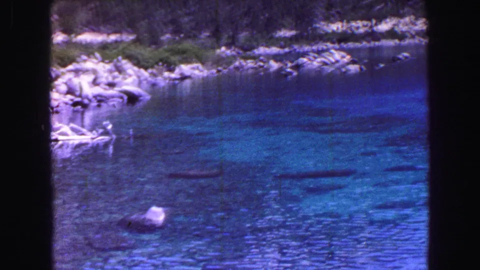 1968 Vancouver 8mm frame showing clear blue water, rounded stones below the surface, rocky shoreline, and foliage at the edge.