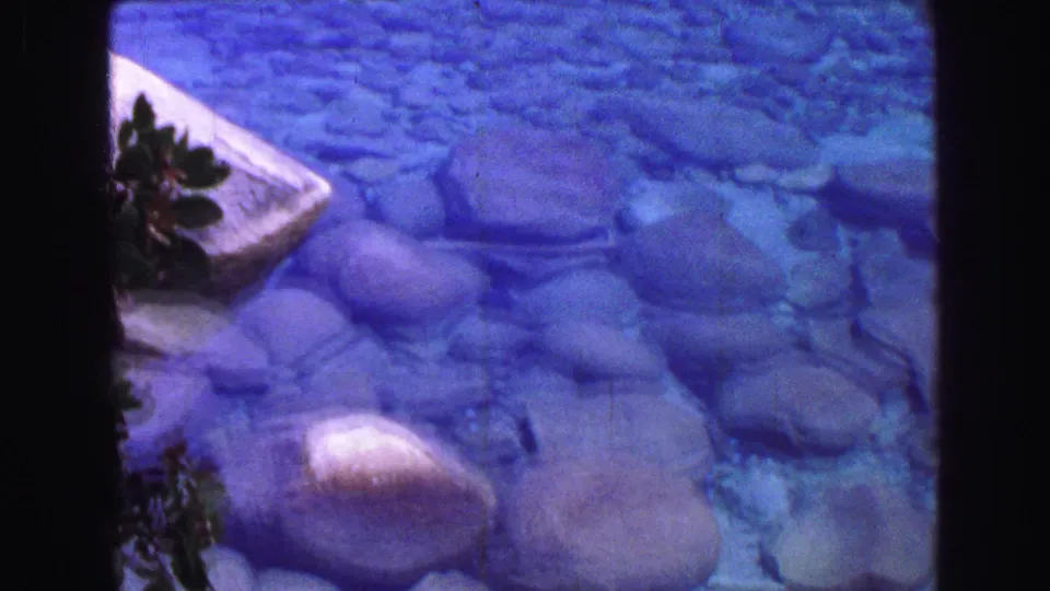 1968 Vancouver frame showing a closer pass over clear blue water and smooth submerged stones near foliage and a pale shoreline edge.
