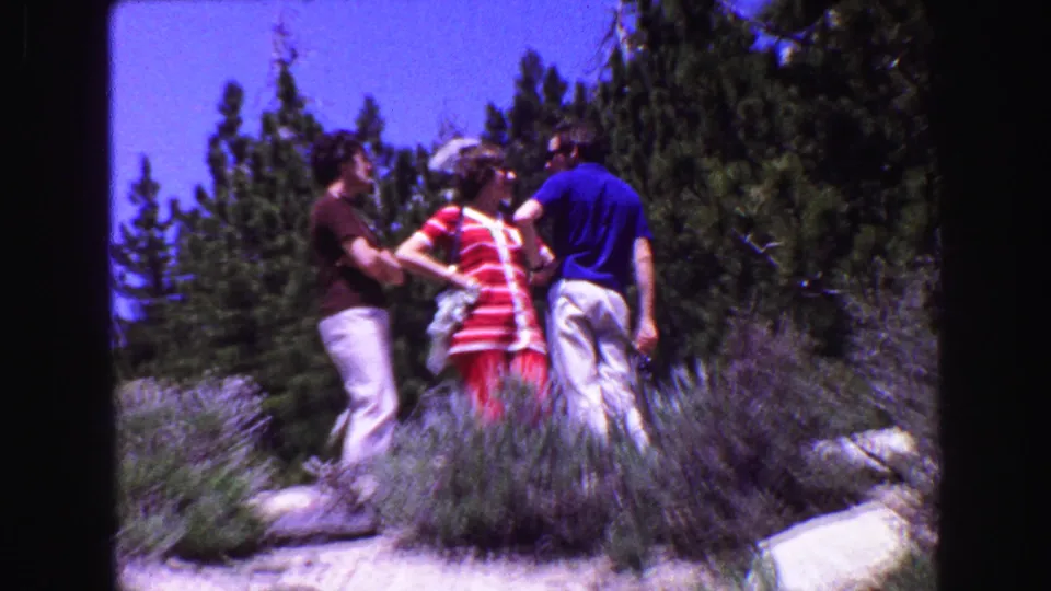 Three adults appear to talk on a rocky, forested hillside in 1968 British Columbia; the woman wears a red and white striped dress.