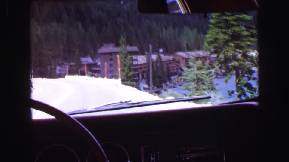 Passenger-side car view in 1968 footage showing a road descending toward rustic lakeside buildings surrounded by evergreen forest.