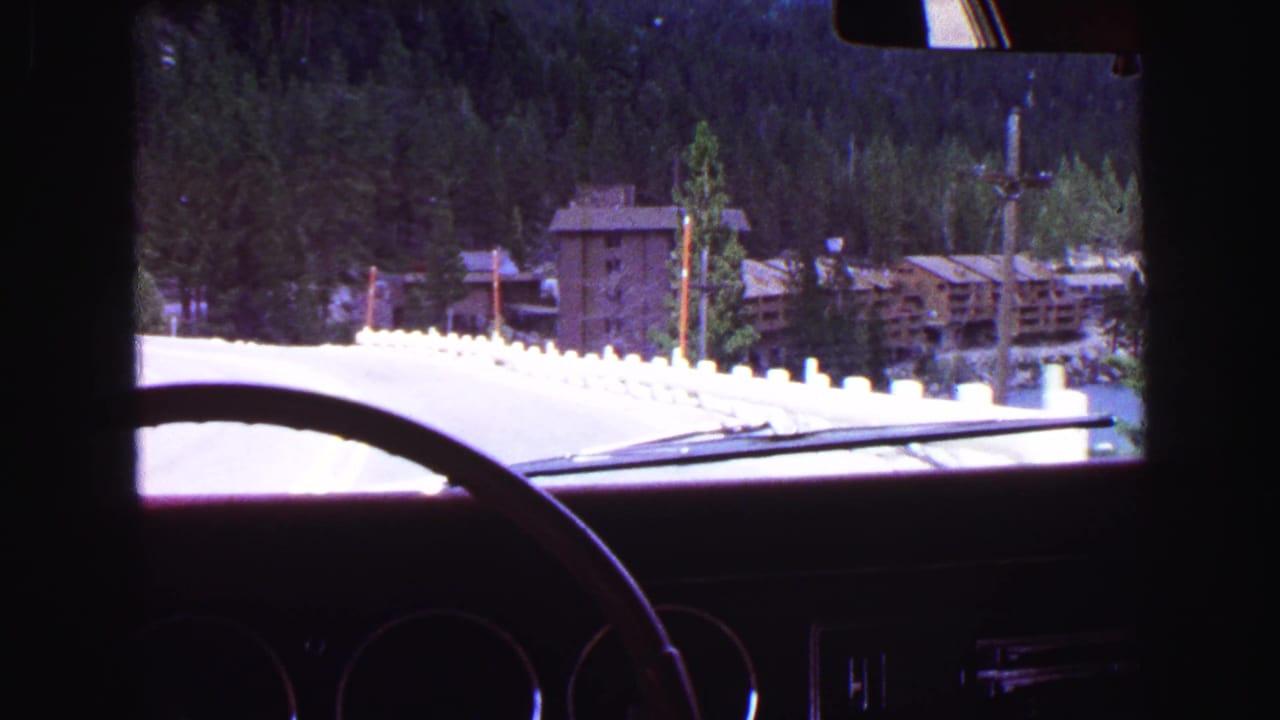 View through a vintage car windshield toward a snowy road, evergreens, and buildings.