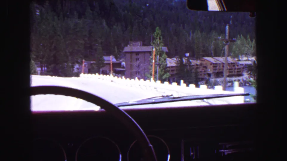1968 8mm view from inside a car on a mountain road near Vancouver, with forest and lodge-like buildings ahead.