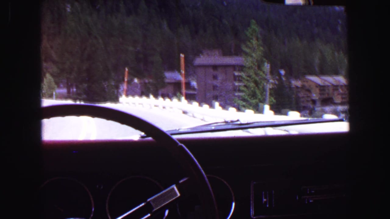 Car interior frames a road curving past forest and rustic buildings.
