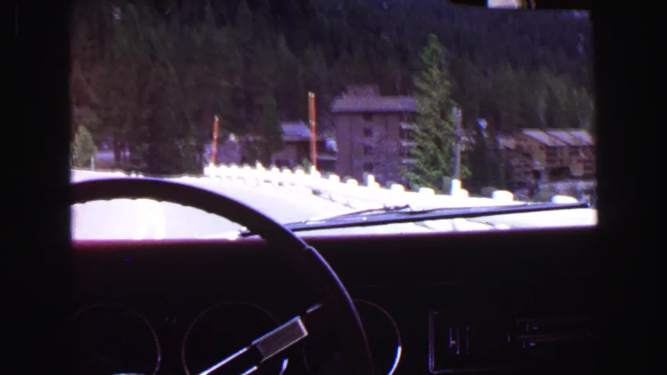 Inside-car view of a 1968 British Columbia mountain drive, with evergreens, roadside posts, and lodge-like buildings visible.