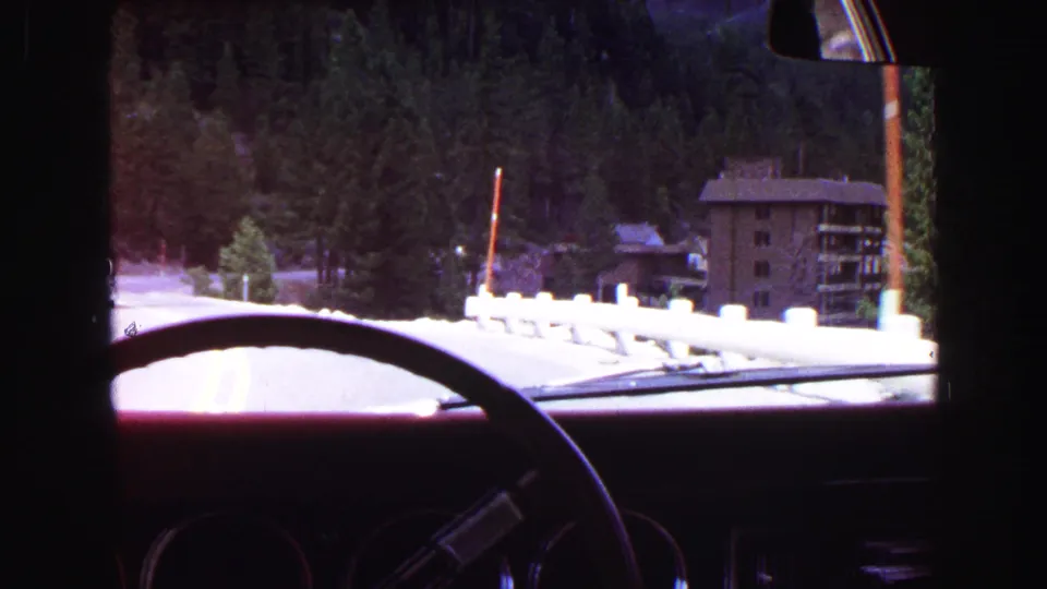 1968 home-movie frame from inside a car, showing a mountain road edge, evergreens, and a multi-story building near Vancouver.
