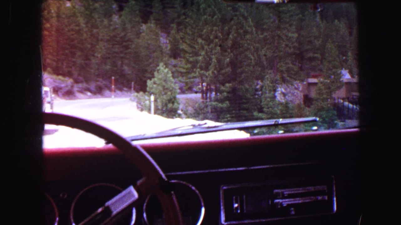 Car dashboard and windshield view of a forest road curving through mountains.