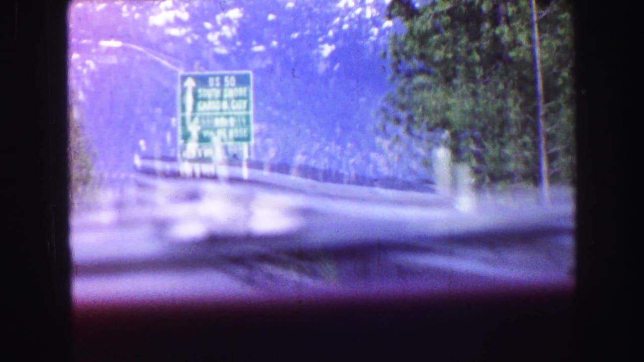 Windshield view approaching a green U.S. 50 direction sign on a forested mountain road, with dashboard glare and trees visible.