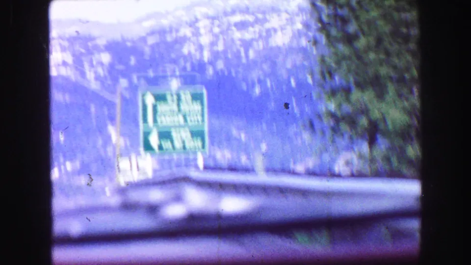 Soft 8mm frame of a green highway sign beside a mountain road, filmed through a windshield from inside a moving car.