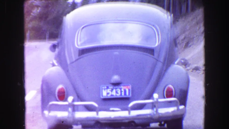 1968 Vancouver-area 8mm frame showing the rear of a blue Volkswagen Beetle driving on a tree-lined road.