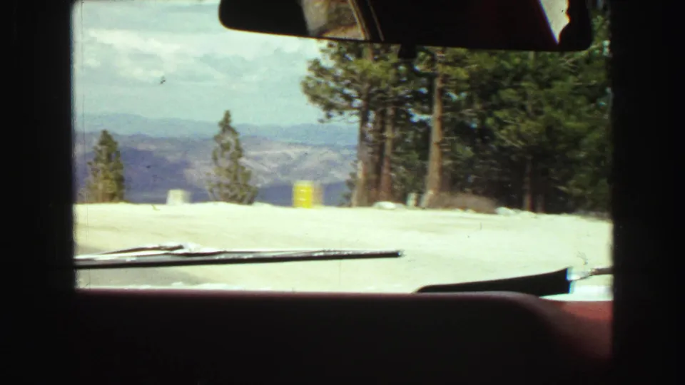 1968 car interior view across a snowy mountain overlook with dashboard, mirror, wiper blade, evergreens, and distant ridges.