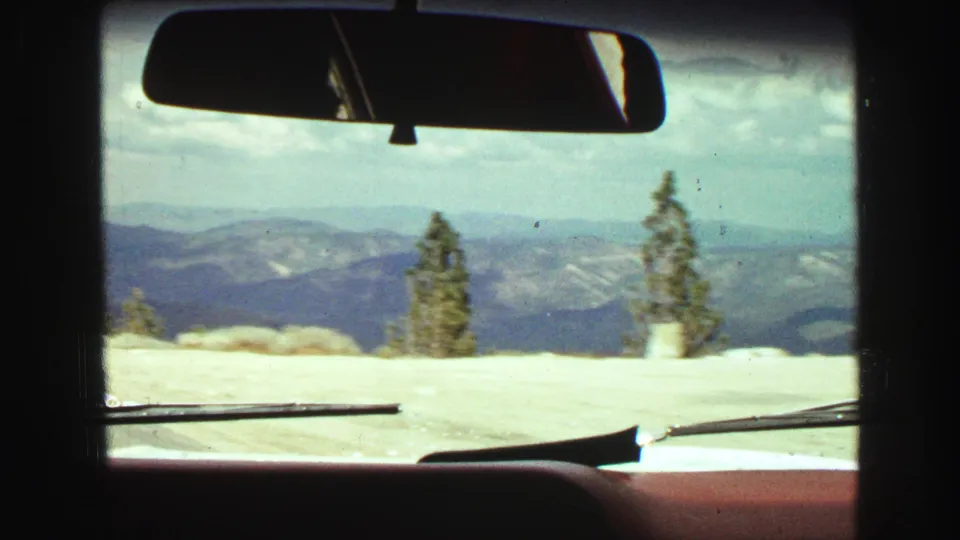 Passenger-shot 1968 8mm frame looking through a car windshield at snow, evergreens, and distant British Columbia mountain ridges.