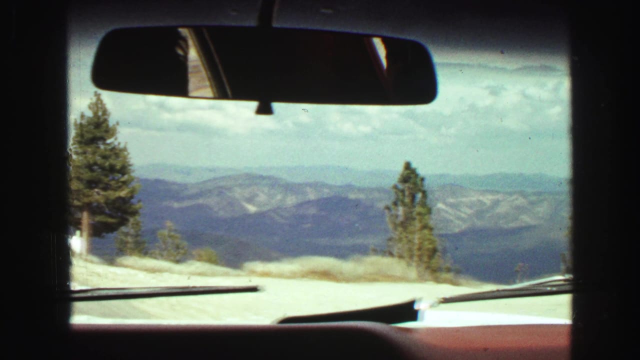 Car windshield view of a mountain road with trees, distant hills, dashboard, and rearview mirror.