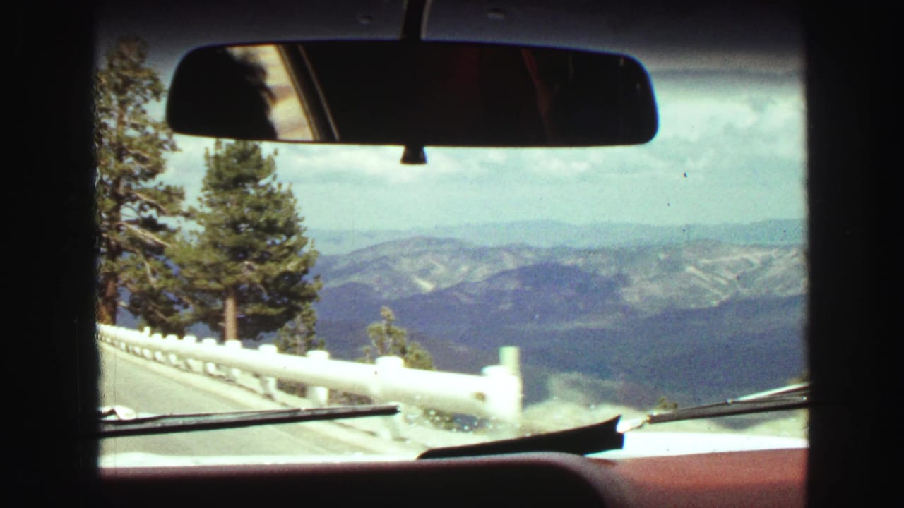 Windshield view with roadside guardrail, evergreen trees, distant mountains, dashboard, and mirror.