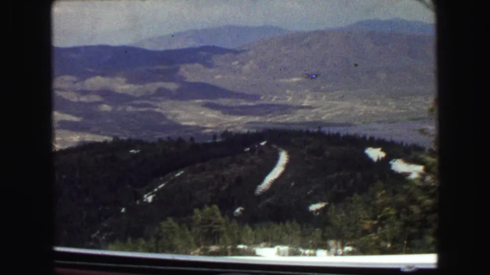 1968 8mm view near Vancouver looking through a car window toward a mountain valley, forested slopes, snow patches, and hazy distant ridges.