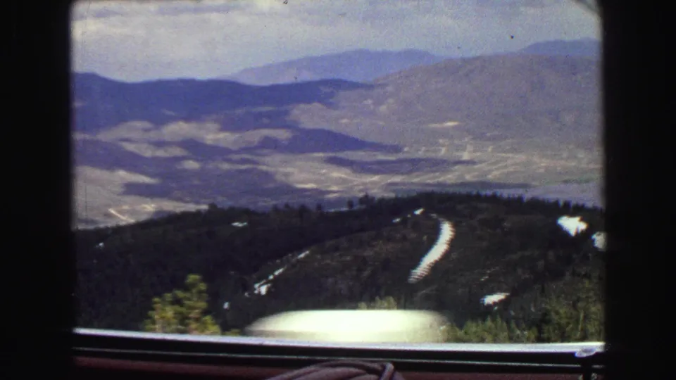 1968 8mm car-window view near Vancouver showing forested mountain slopes, bright snow patches, a distant valley, ridgelines, and the car hood or dashboard edge.