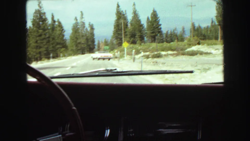 Windshield view of a forested mountain road with another car ahead, filmed on 8mm near Vancouver, British Columbia in 1968.