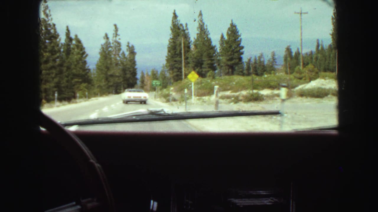 Windshield view of a mountain road with a white sedan ahead and evergreen trees along the roadside.