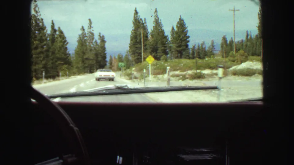 1968 Vancouver-area home-movie view from inside a car following a white sedan on a two-lane mountain road lined with evergreens.