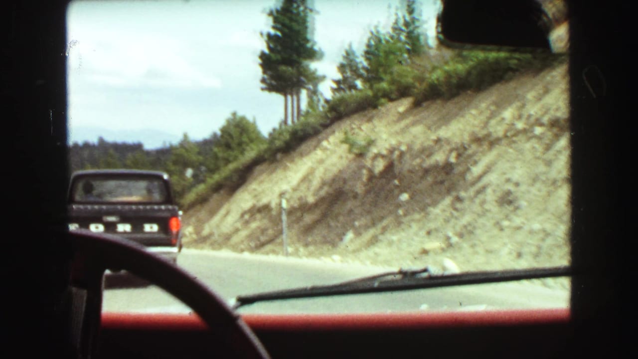 View through a car windshield following a black pickup truck beside a cut hillside.