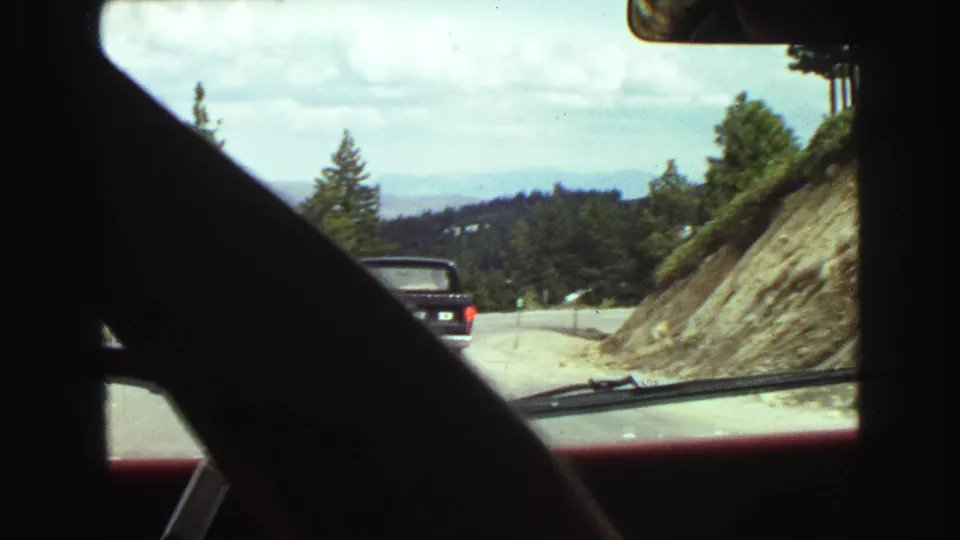 1968 in-car home-movie frame showing a black pickup truck on a mountain road with hillside and evergreens nearby.