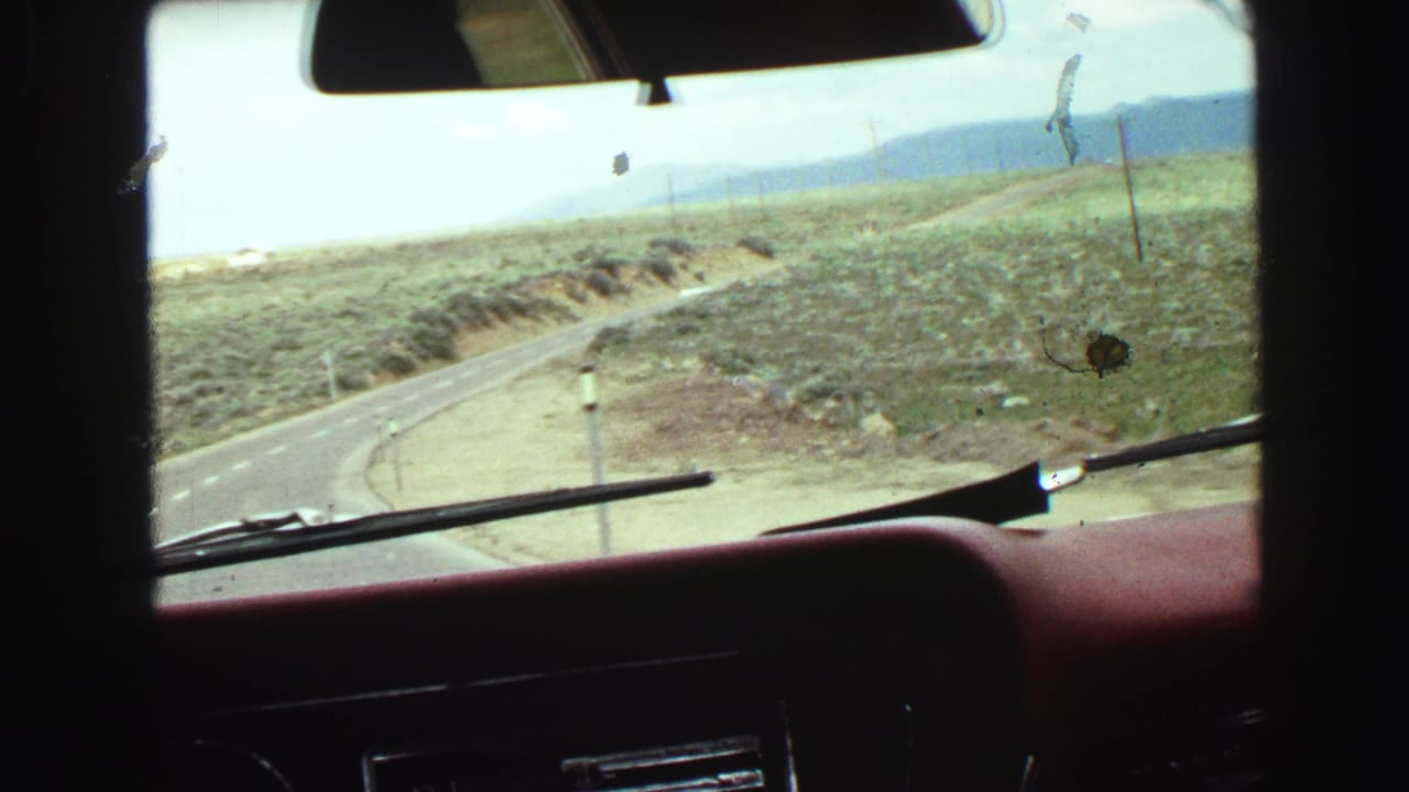 Windshield view of a curving road through dry open terrain and distant hills.